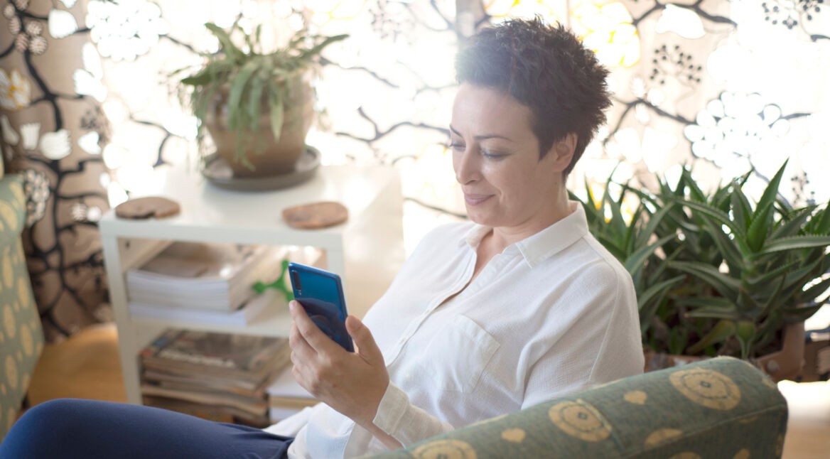 Eine Frau sitzt mit ihrem Smartphone in einem Sessel, umgeben von Pflanzen