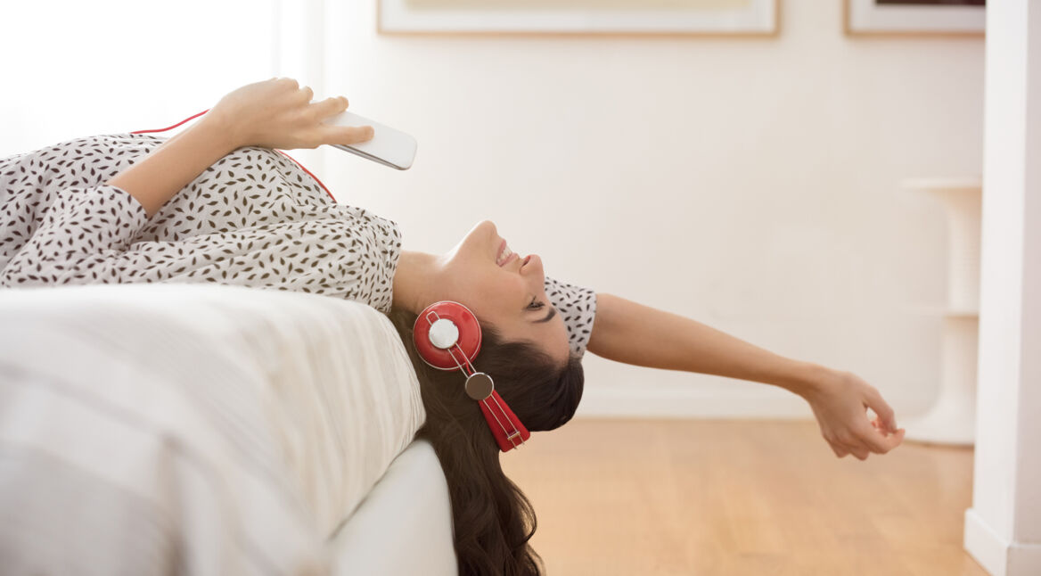 Eine Frau mit roten Kopfhörern liegt auf dem Bett und hört etwas auf ihrem Handy.