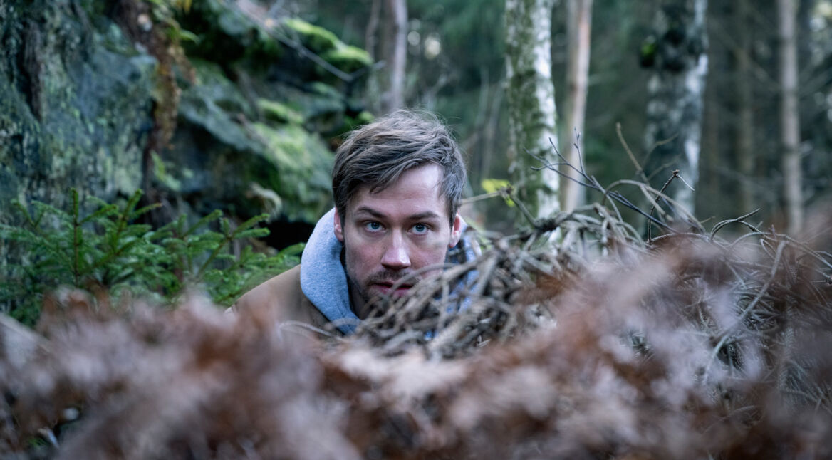 Roman (David Kross) versteckt sich im Wald hinter Ast-Gestrüpp.