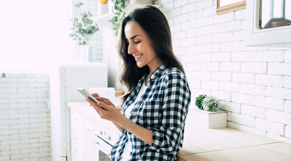 Lächelnde Frau schaut in der Küche aufs Smartphone