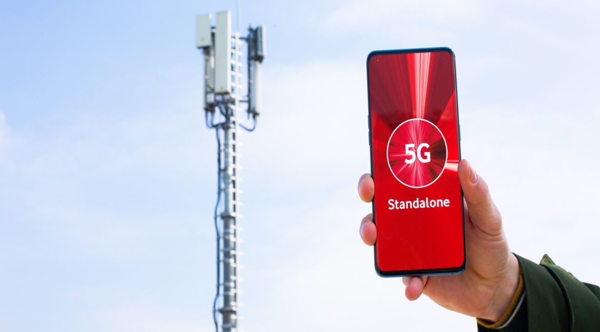 Ein Smartphone, dass auf dem Bildschirm "5G Standalone" anzeigt, wird in die Luft gehalten. Im Hintergrund ist ein Mobilfunkmast zu sehen.