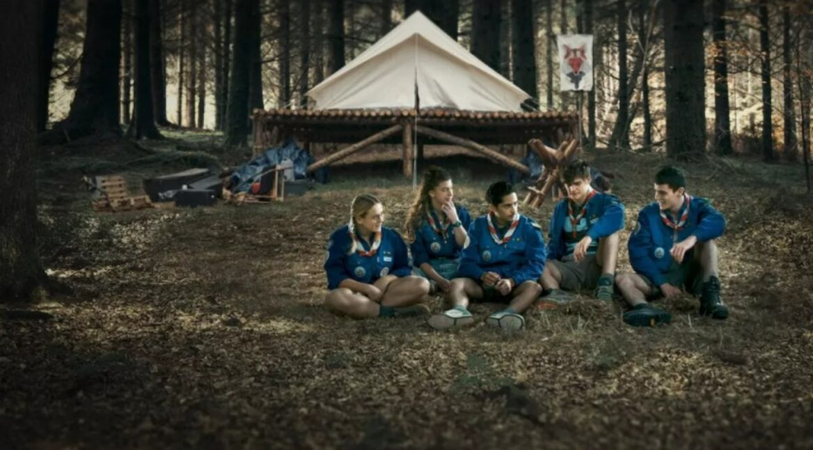 Fünf junge Pfadfinder:inen sitzen vor einem Zelt im Wald.