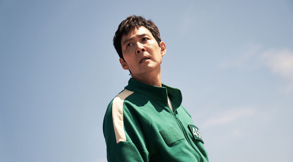 Auf dem Bild aus "Squid Game Staffel 2" ist der Charakter Seong Gi-hun, gespielt von Lee Jung-jae, zu sehen. Er trägt eine grüne Trainingsjacke mit der Nummer 456 und schaut angespannt in die Ferne. Der Himmel im Hintergrund ist klar und blau, was die ernste und nachdenkliche Stimmung des Moments unterstreicht.