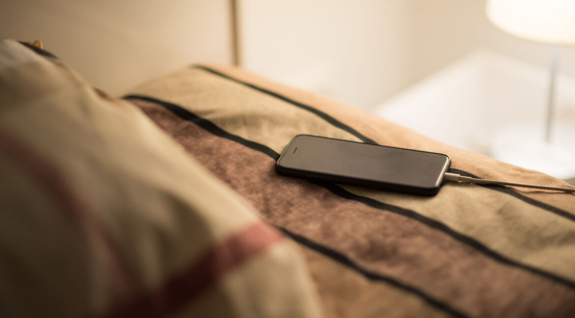 Ein iPhone liegt auf einem Bett und lädt per Kabel auf.