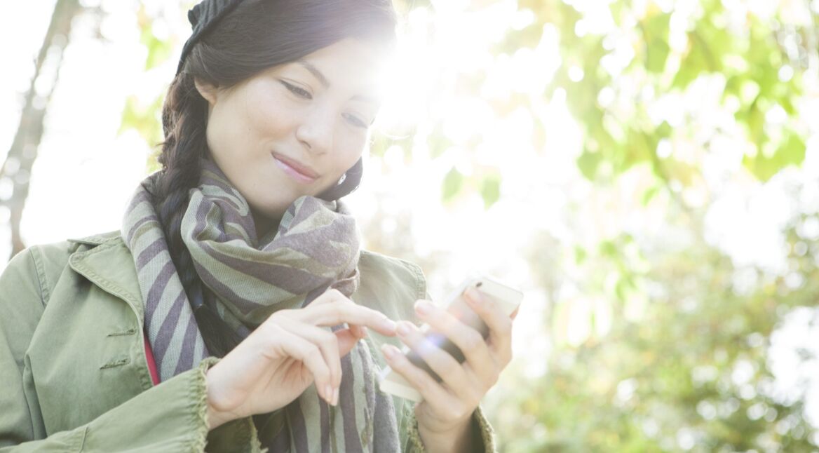 Frau mit Sonne und Baum im Rücken guckt lächelnd auf ihr iPhone