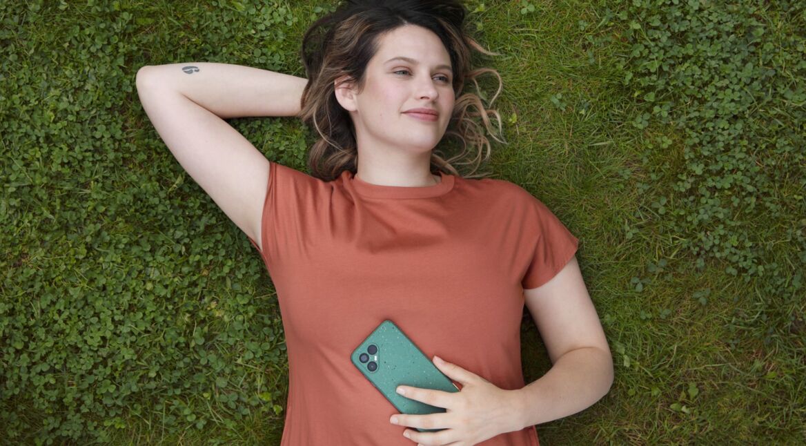 Lächelnde Frau liegt auf grüner Wiese und hält Fairphone 4