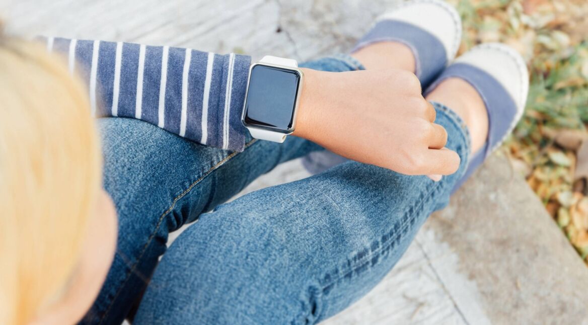 Blonde Frau sitzt auf Brüstung und schaut auf Apple Watch am Handgelenk