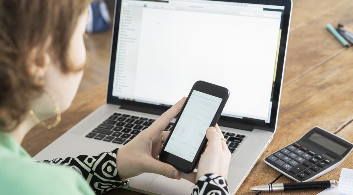 Frau sitzt vor Laptop und hält Smartphone in beiden Händen