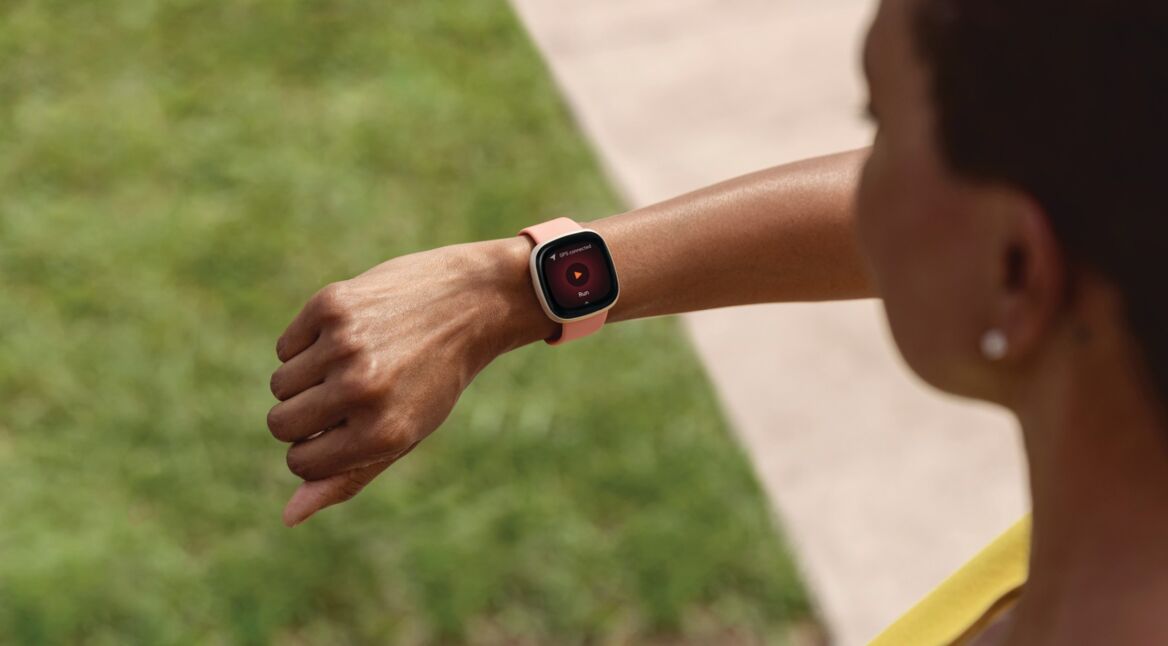 Blick über Schulter: Frau schaut draußen auf ihre Smartwatch Fitbit Versa 3 am rechten Handgelenk.