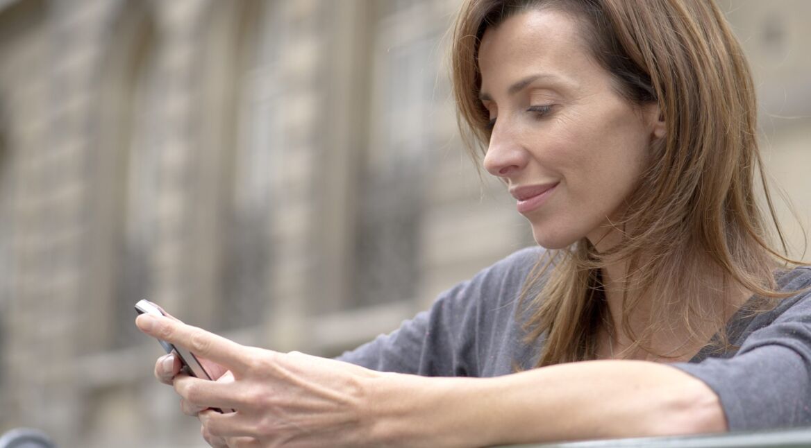 Frau steht draußen und schaut lächelnd auf ihr Smartphone