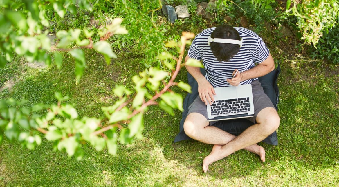 Ein Mann mit Kopfhörer und Laptop sitzt im Grünen.