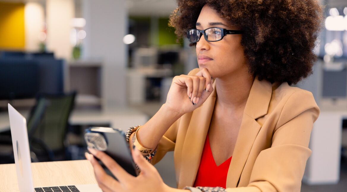 Frau sitzt nachdenklich vor ihrem MacBook und hält ein iPhone in der Hand