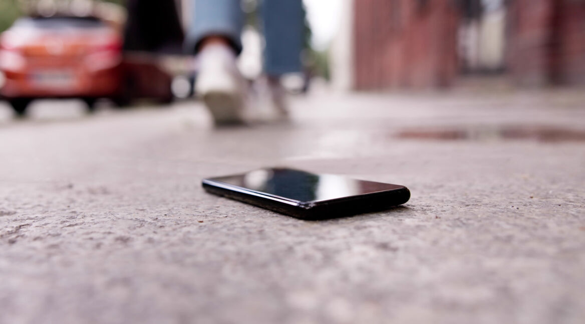 Auf dem Bild zum Thema Handy verloren oder gestohlen ist ein schwarzes Smartphone zu sehen, das unbeachtet auf einem grauen Gehweg liegt. Im unscharfen Hintergrund entfernt sich eine Person in Jeans und weißen Sneakern, links parkt ein rotes Auto, rechts ragt eine Backsteinfassade auf. Die bodennahe Perspektive und das gedämpfte Tageslicht vermitteln ein Gefühl von Verlust und Unachtsamkeit.