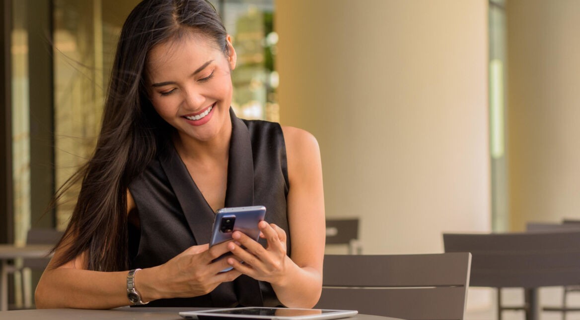 Asiatische Frau sitzt draußen im Cafè und guckt auf ihr Handy