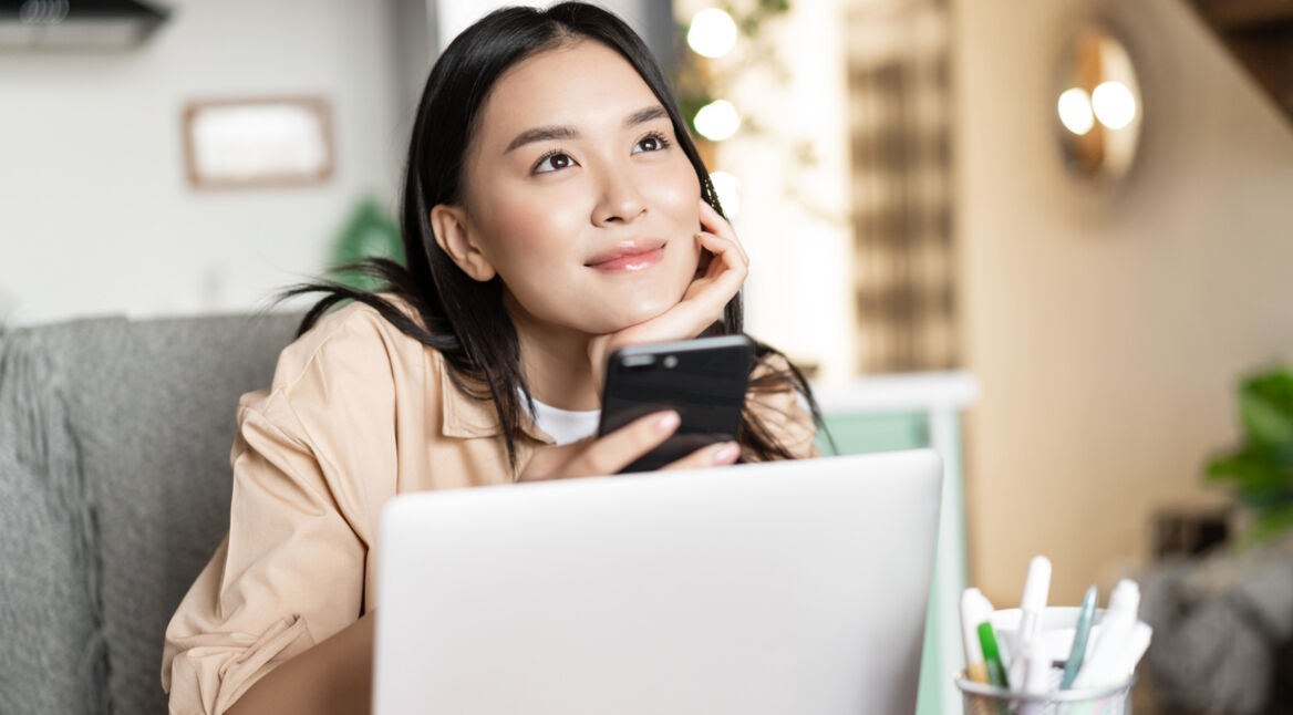 Frau vor Laptop mit Smartphone in der Hand schaut nachdenklich in die Ferne