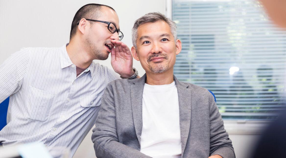 Das Bild zeigt zwei Männer. Der linke Mann flüstert dem rechten Mann etwas ins Ohr.