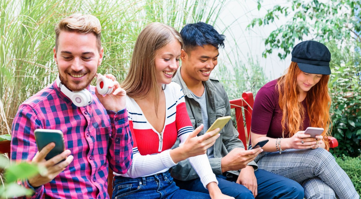 Vier junge Menschen sitzen an einem Tech und gucken lachend auf ihre Handys