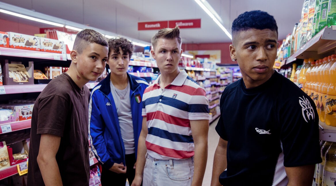 Lukas (Levy Rico Arcos), Gino (Rafael Klein-Heßling), Julius (Vincent Wiemer) und Sanchez (Aaron Maldonado-Morales) stehen in einem Supermarkt und besorgen, was sie für ihren Abend brauchen.
