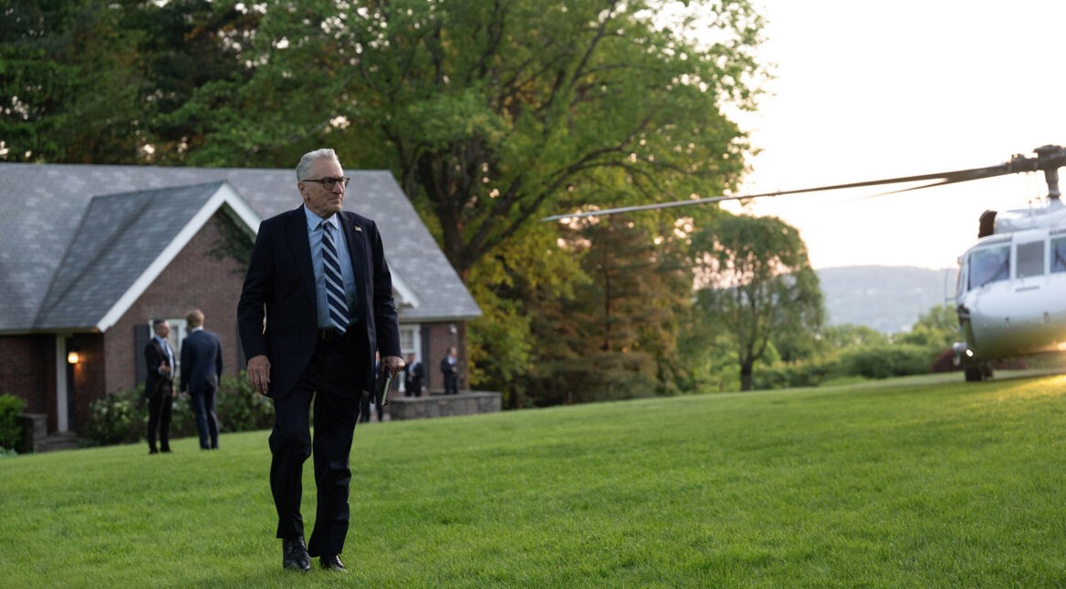 Auf dem Bild zur Serie Zero Day mit Robert De Niro ist Robert De Niro in einer eleganten schwarzen Anzug-Krawatten-Kombination zu sehen. Er geht über eine gepflegte Rasenfläche vor einem Backsteinhaus, während im Hintergrund ein weißer Helikopter steht. Die Szene wirkt angespannt und professionell, unterstützt durch die Anwesenheit von Sicherheitskräften im Hintergrund.