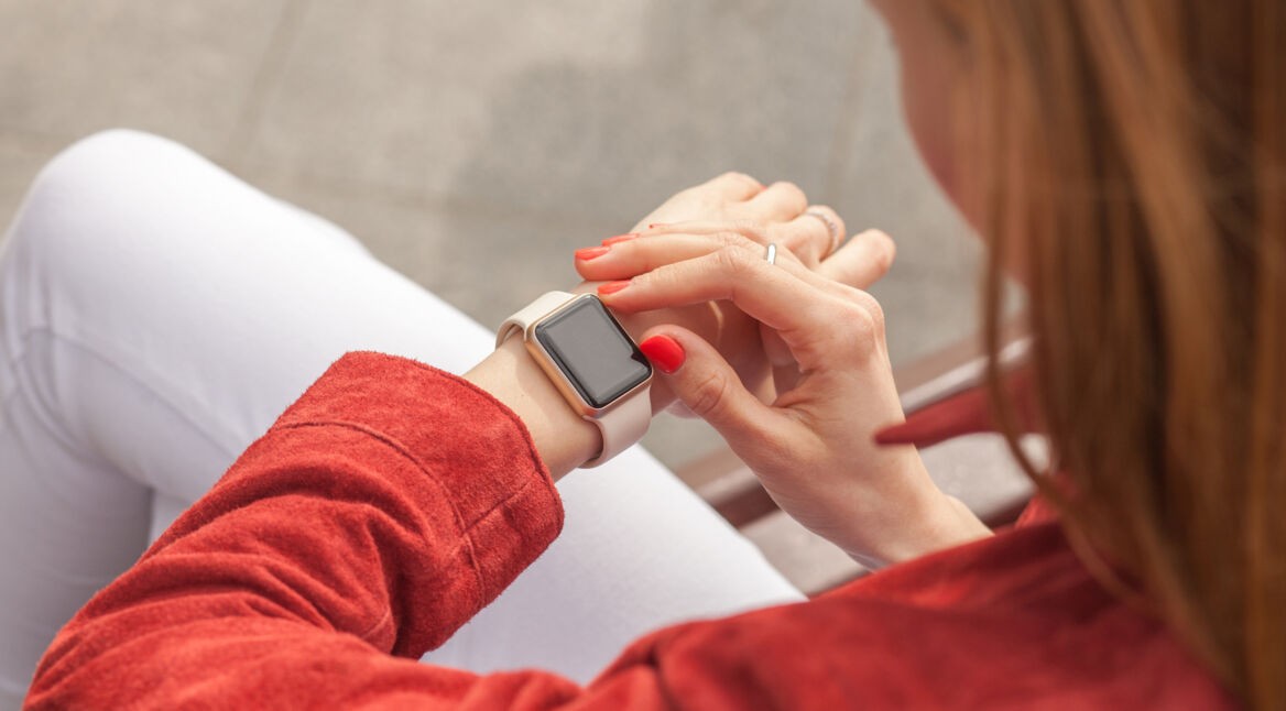 Auf dem Bild zur Smartwatch für Damen ist eine Frau in roter Kleidung zu sehen, die entspannt auf ihr Handgelenk blickt und eine moderne Smartwatch bedient. Die helle Uhr mit rechteckigem Display steht im Mittelpunkt der Szene. Durch die Nahaufnahme wirkt das Motiv stilvoll und modern, während die warme Farbgebung eine ruhige und elegante Stimmung vermittelt.