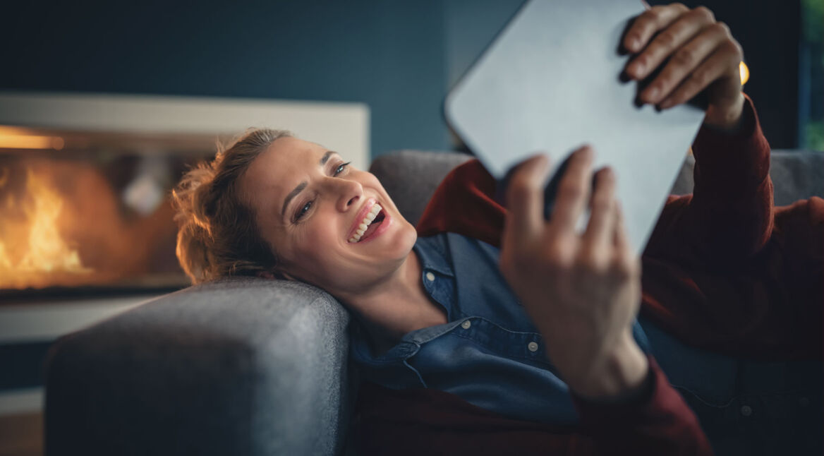 Lachende Frau liegt auf dem Sofa mit ihrem Tablet in der Hand