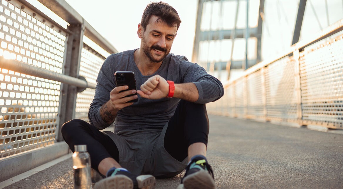 Mann sitzt auf dem Boden, macht Pause vom Sport und schaut lächelnd auf seine Smartwatch und sein Handy