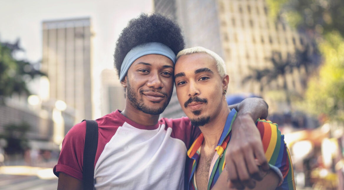 Auf dem Bild zur CSD Vodafone Kampagne umarmen sich zwei Männer und schauen selbstbewusst in die Kamera. Einer trägt ein Hemd in Regenbogenfarben, das Symbol für Pride und Vielfalt. Die Szene spielt in einer belebten Innenstadt mit Hochhäusern und sommerlicher Atmosphäre – ein Zeichen für Zusammenhalt, Sichtbarkeit und Offenheit.