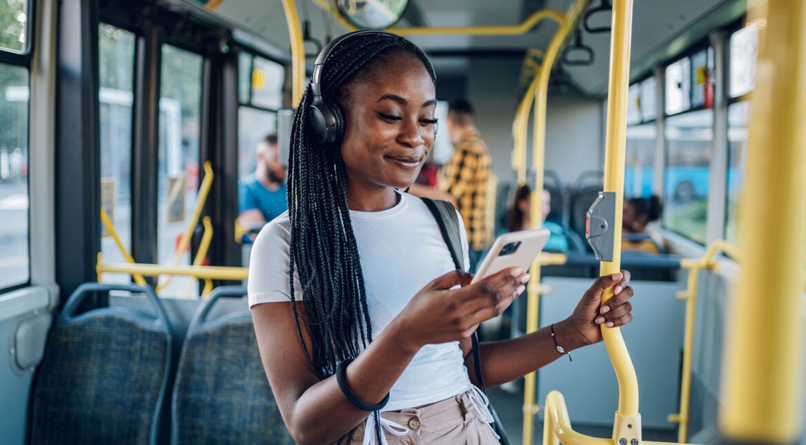 Junge Frau im Bus hört Musik und schaut lächelnd auf ihr Handy