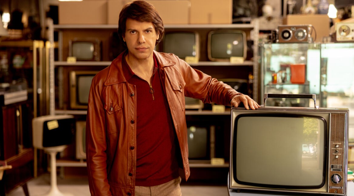 Ein Mann in brauner Lederjacke steht mit der Hand auf einem Fernseher in einem Laden.