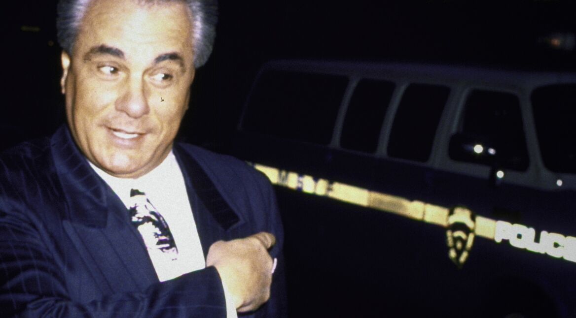 John Gotti: Ein Anzug tragender Mann mit zurückgekämmtem, grauem Haar, deutet auf einen Polizeiwagen.