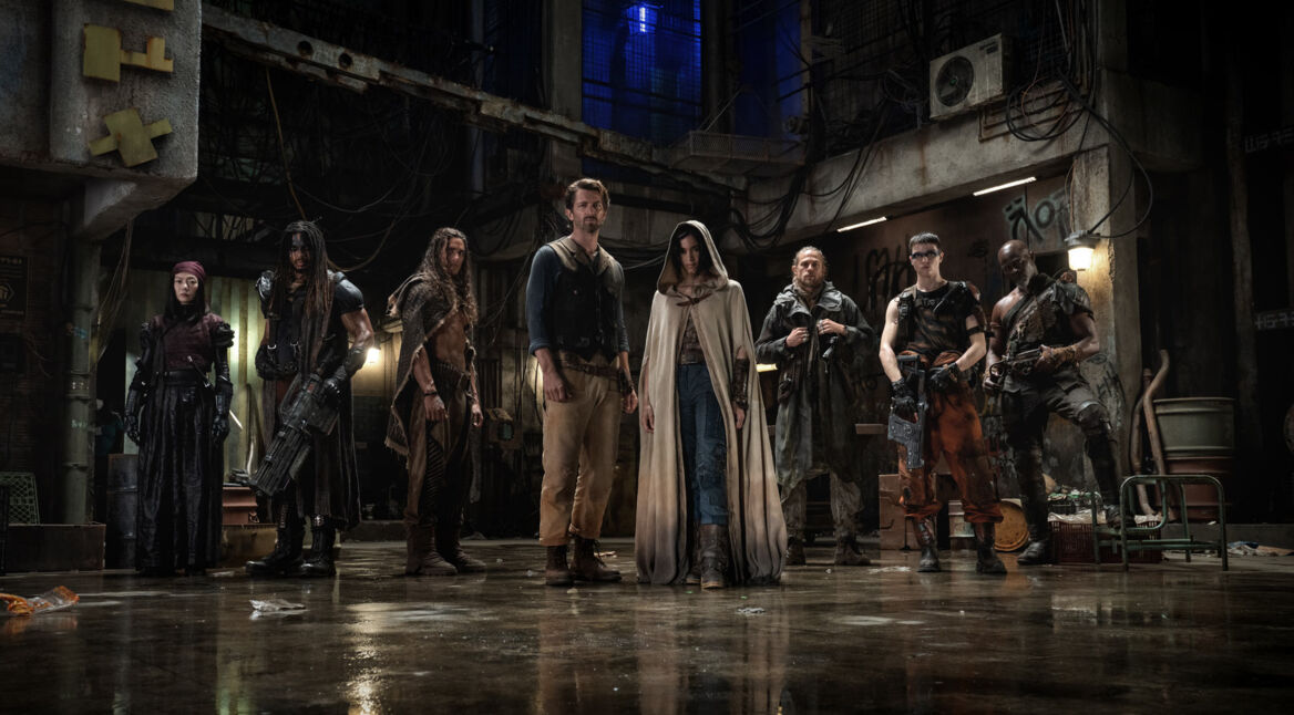 Der Rebel Moon Cast steht nebeneinander aufgereiht in einer alten Fabrikhalle