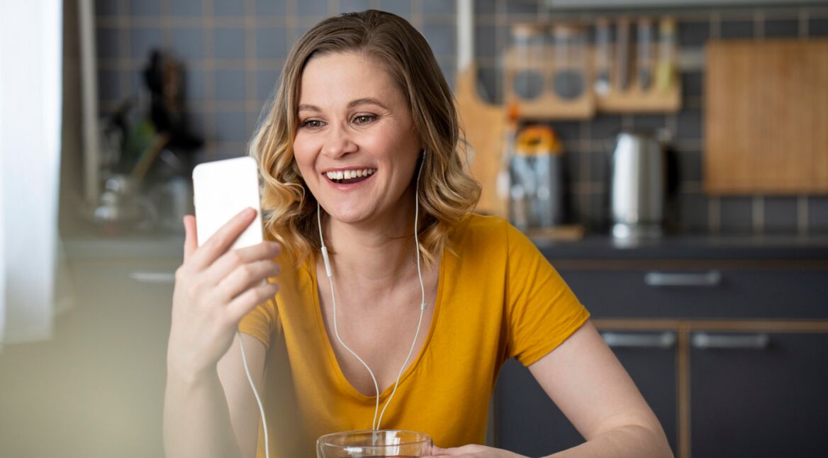 Eine junge Frau sitzt in einer Küche, schaut in ihr Smartphone und lächelt dabei.