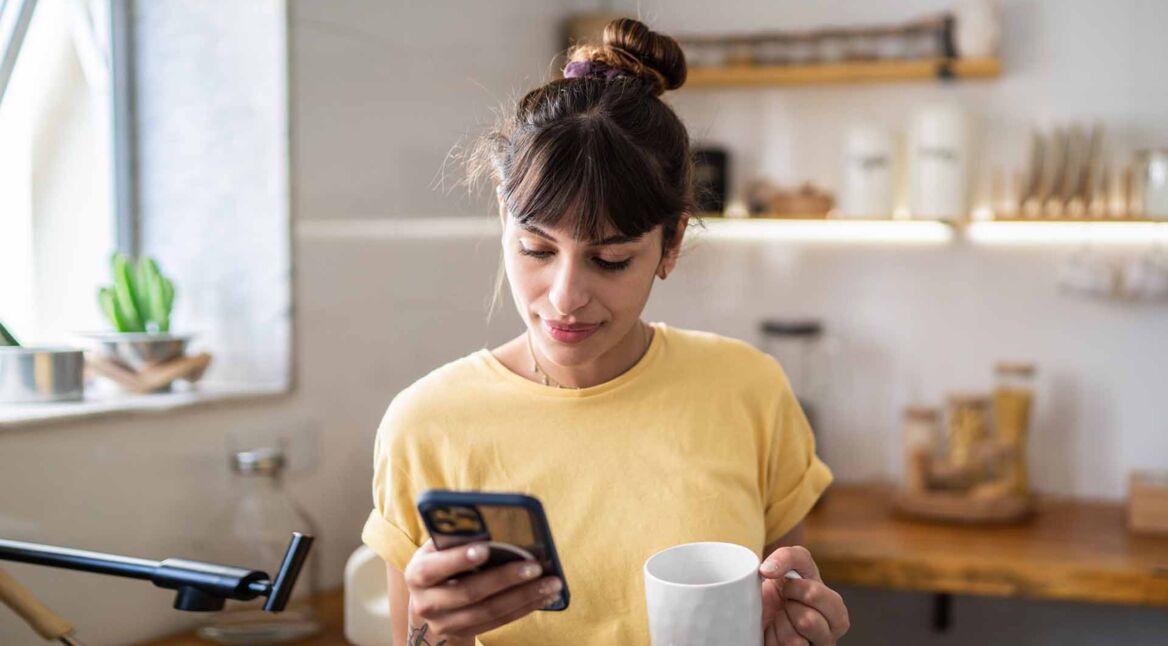Junge Frau, die das Handy benutzt, während sie zu Hause Kaffee oder Tee trinkt
