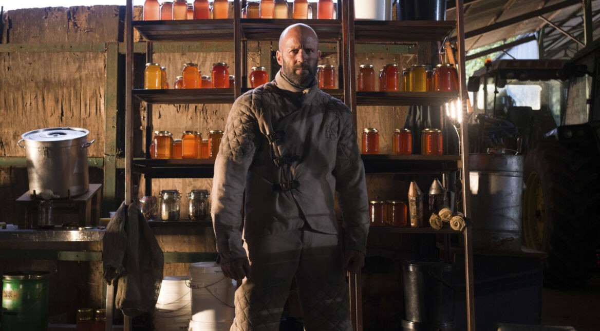 The Beekeeper streamen: Ein Mann im Imker-Anzug steht mit entschlossenem Blick vor einem Regal voller Honiggläser.