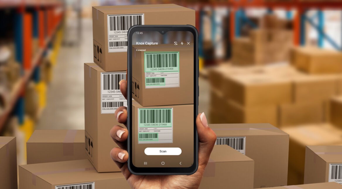 POV: Rechte Hand hält das robuiste Handy Galaxy Xcover7 in einer Lagerhalee vor einem Stapel Kartons