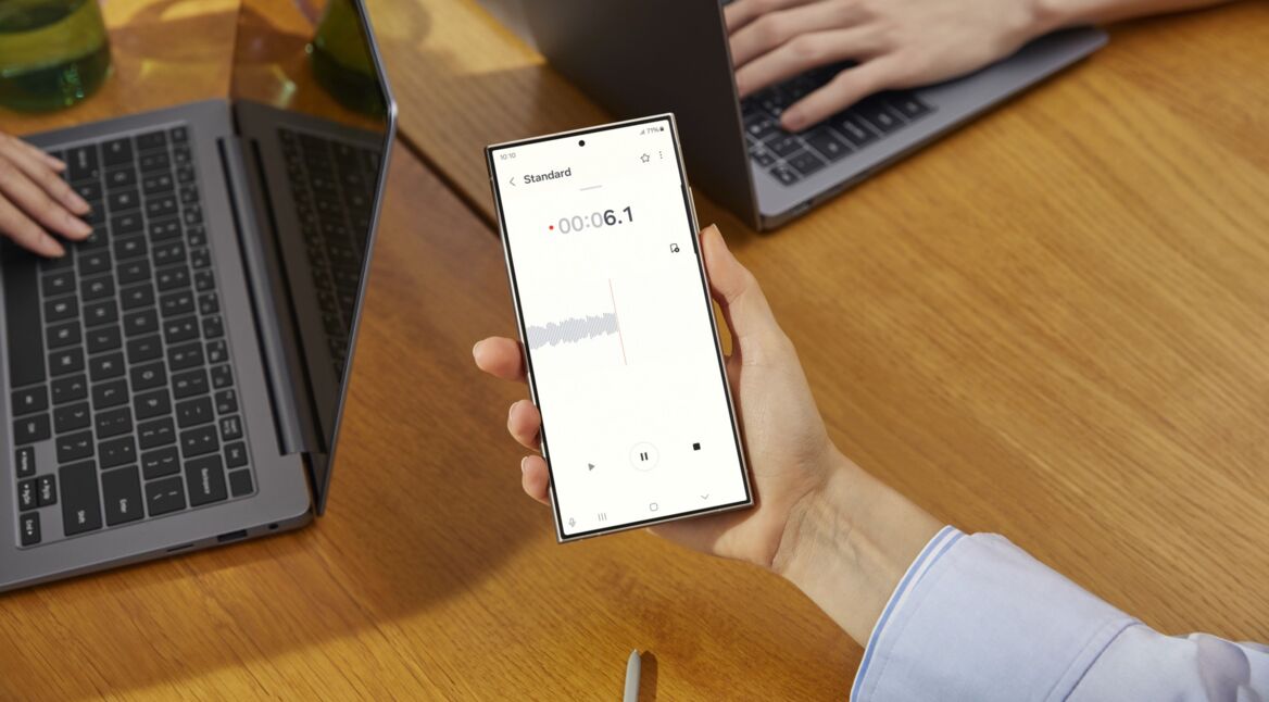 Eine Hand hält das Samsung Galaxy S24 Ultra über einem Schreibtisch, dahinter zwei Notebooks.