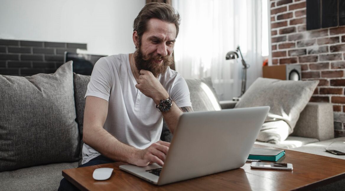 Ein Mann sitzt auf der Couch und nutzt dabei einen Laptop auf dem Tisch vor sich.