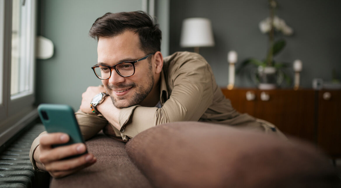 Ein Mann mit Brille liegt lächelnd auf einer Couch und hält ein Smartphone in seinen Händen und nutzt eine der besten kostenlosen Dating-Apps