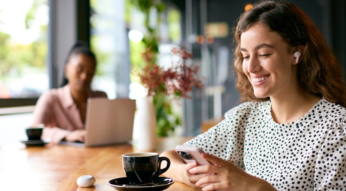 Frau sitzt lächelnd an einem Tisch mit einer Tasse vor sich, hat Handy in der Hand und Kopfhörer im Ohr