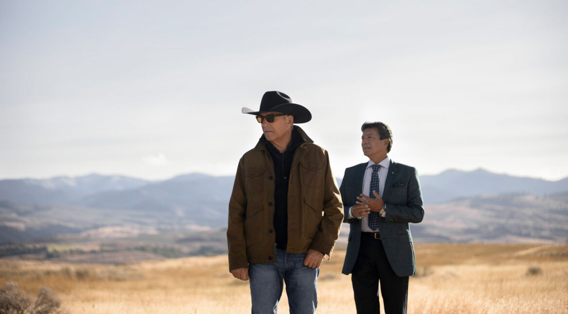 Auf dem Bild zum Faktenartikel über die Serie Yellowstone stehen Kevin Costner und Gil Birmingham in einer weiten Landschaft mit goldgelbem Gras und Bergen im Hintergrund. Kevin Costner trägt eine braune Jacke, Jeans, Sonnenbrille und einen schwarzen Cowboyhut, während Gil Birmingham einen eleganten grauen Anzug mit Krawatte trägt. Die Szene wirkt ruhig und nachdenklich, betont durch die offene Natur.