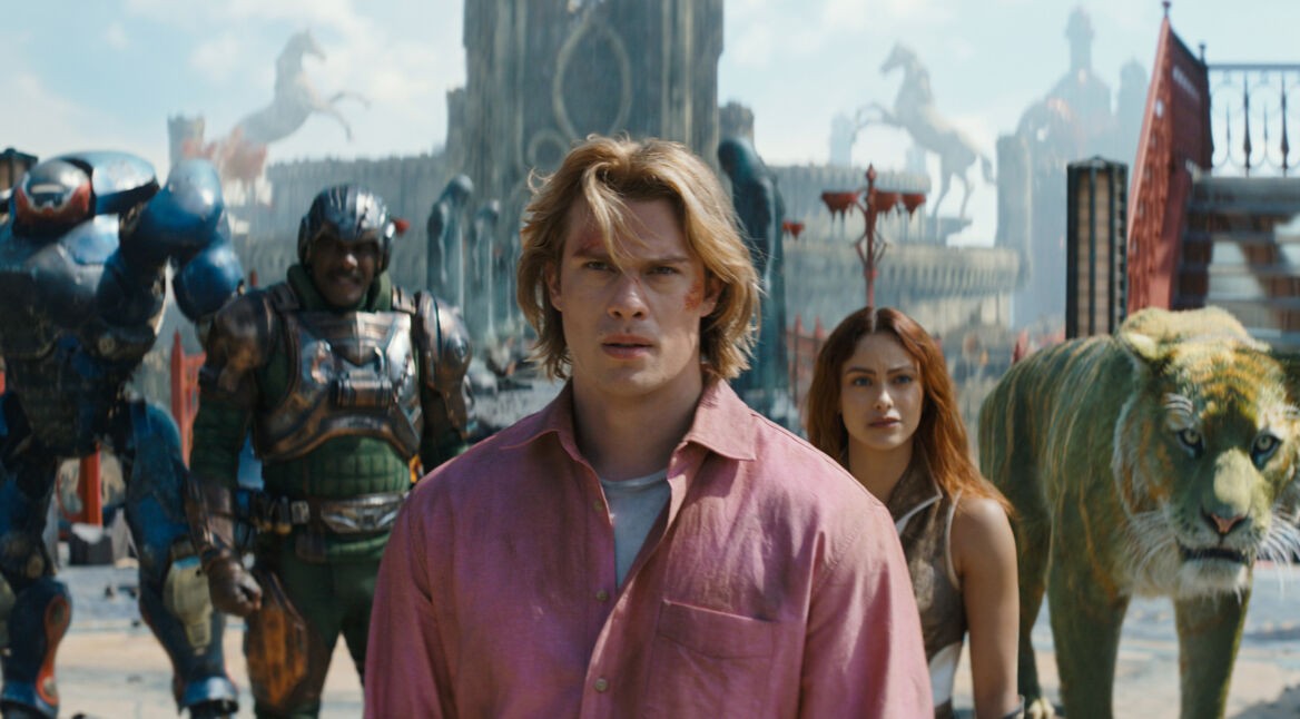 Im Bild zum Masters of the Universe neuer Film ist Adam, gespielt von Nicholas Galitzine, im Vordergrund mit entschlossenem Blick zu sehen. Hinter ihm stehen Teela (Camila Mendes), Man-At-Arms (Idris Elba), Roboto (Kristen Wiig) und der grüne Tiger Cringer. Die Szene wirkt episch und angespannt, mit einer fantasievollen, mittelalterlich-futuristischen Kulisse im Hintergrund.