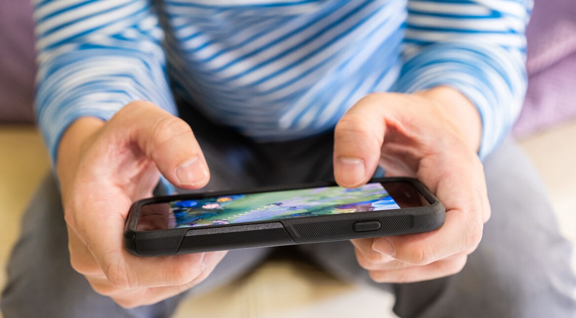 Auf dem Bild zu den besten Handyspielen 2025 hält eine Person mit blau-weiß gestreiftem Shirt ein Smartphone in beiden Händen. Auf dem Bildschirm ist ein farbenfrohes Handyspiel mit grüner Landschaft zu sehen. Die Szene vermittelt eine entspannte Atmosphäre und hebt die Popularität von Mobile Gaming hervor.