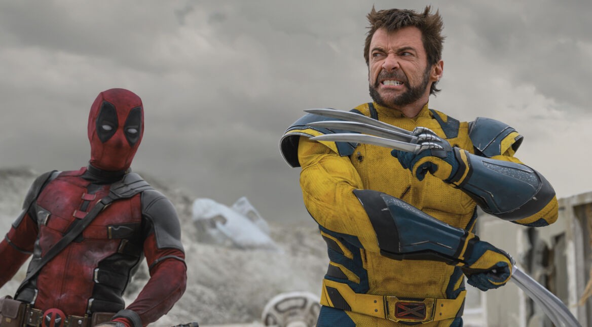 Auf dem Bild zu "Deadpool & Wolverine Ende erklärt" sind die Charaktere Deadpool und Wolverine in einer intensiven Szene zu sehen. Links steht Deadpool, gespielt von Ryan Reynolds, in seinem roten Anzug mit schwarzen Details, ernst und aufmerksam. Rechts von ihm steht Wolverine, dargestellt von Hugh Jackman, in einem gelben Anzug mit blauen Akzenten und ausgefahrenen Adamantium-Krallen, bereit zum Kampf. Im Hintergrund ist eine karge, graue Landschaft unter einem bewölkten Himmel zu sehen, was die Spannung der Szene unterstreicht.
