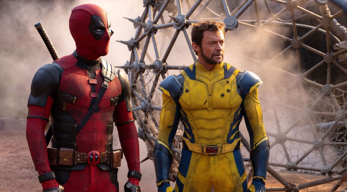 Das Bild zur Deadpool & Wolverine-Kritik zeigt Ryan Reynolds als Deadpool und Hugh Jackman als Wolverine. Beide Charaktere stehen nebeneinander vor einer rauchigen, futuristischen Kulisse, die möglicherweise an ein Schlachtfeld oder eine industrielle Umgebung erinnert. Deadpool (Ryan Reynolds) trägt seinen typischen rot-schwarzen Anzug mit mehreren Taschen und Gürteln sowie sein charakteristisches Schwert auf dem Rücken. Wolverine (Hugh Jackman) ist in einem gelb-blauen Anzug zu sehen, der stark an das klassische Comic-Kostüm des Charakters erinnert. Der Anzug betont seine muskulöse Statur und ist mit einigen metallischen Akzenten versehen. Beide wirken entschlossen und bereit für die nächste Herausforderung, was durch ihre angespannten und konzentrierten Gesichtsausdrücke unterstrichen wird.