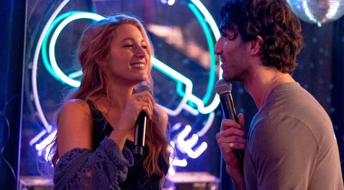 Ein Szenenbild aus dem Nur noch ein einziges Mal-Film zeigt Blake Lively in der linken Bildhälfte als Lily Bloom und Justin Baldony in der rechten Bildhälfte als Ryle. Die beiden schauen sich lächelnd an und haben jeweils ein Mikrofon in der Hand. Im Hintergrund leuchtet ein Karaoke-Symbol in Blau und Lila. Am oberen rechten Bildrand sieht man eine Diskokugel.