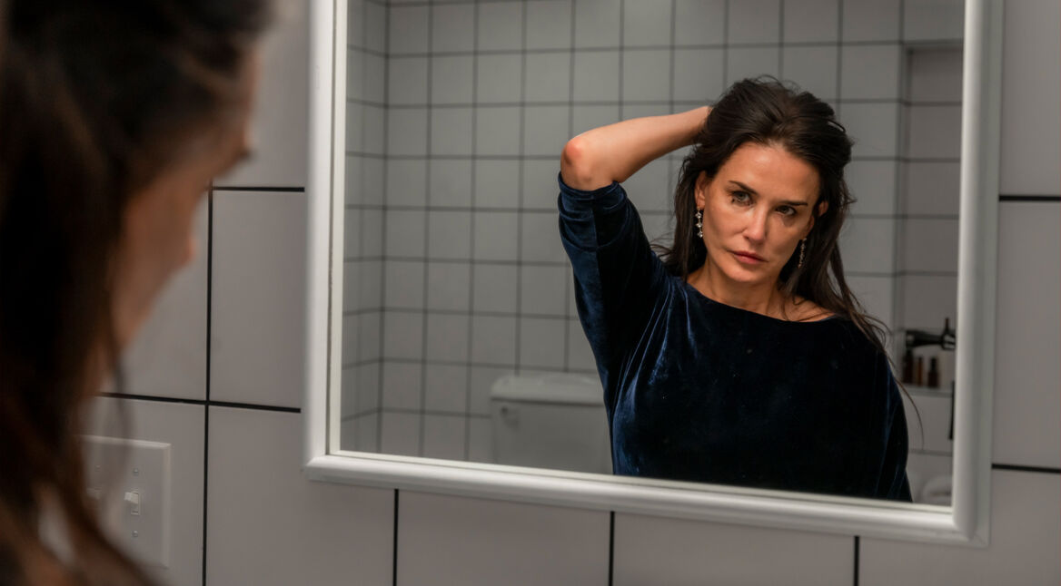 Demi Moore steht in The Substance als Elisabeth Sparkle vor dem Badezimmerspiegel. Am linken Bildrand ist verschwommen ihr Gesicht von hinten-rechts zu erkennen sowie ihre Haare. Zentral im Bild sieht man ihr Spiegelbild. Sie scheint geweint zu haben. Ihre Maskara ist unter den Augen verwischt. Sie hält sich die langen, dunklen Haare mit der linken Hand am Hinterkopf aus dem Gesicht und schaut kritisch ihr eigenes Gesicht im Spiegel an. Sie trägt ein samtig-blaues Oberteil und lange Ohrringe. Hinter ihr sind weiße, monotone, quadratische Fliesen der Badezimmerwand zu sehen.
