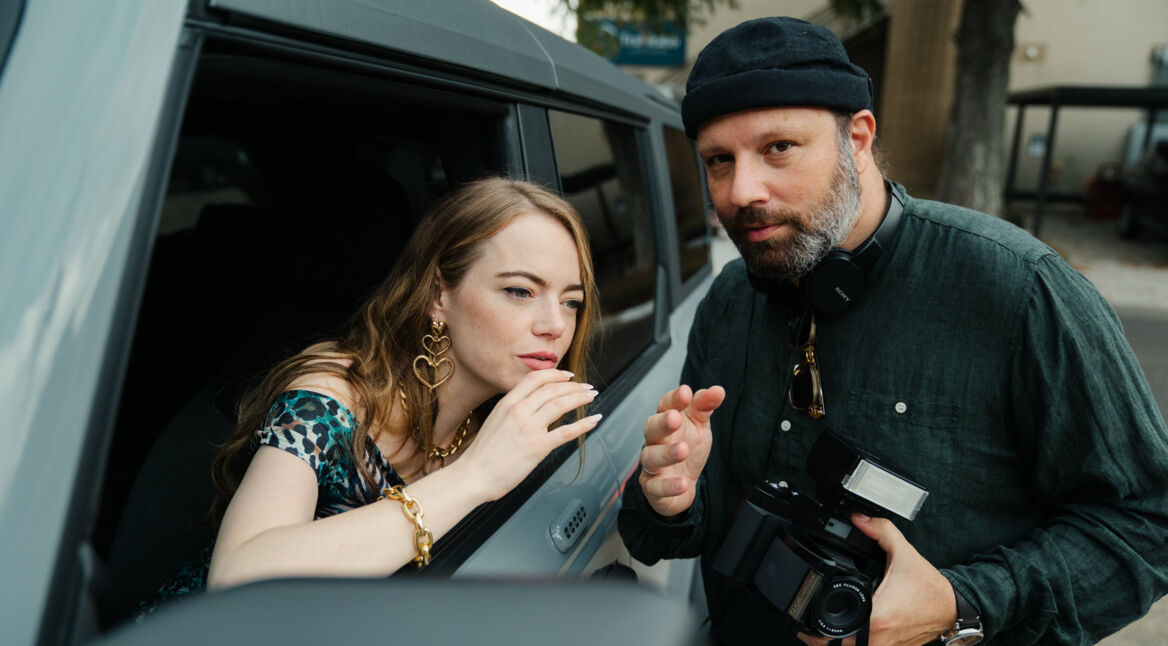 Das Bild auf Yorgos Lanthimos Film-Ranking zeigt Yorgos Lanthimos und Emma Stone in einer Filmszene. Emma Stone schaut konzentriert aus einem Autofenster, während Lanthimos ihr etwas erklärt. Er trägt Kopfhörer um den Hals und hält eine Kamera. Die Szene wirkt professionell und zeigt eine enge Zusammenarbeit zwischen Regisseur und Schauspielerin.