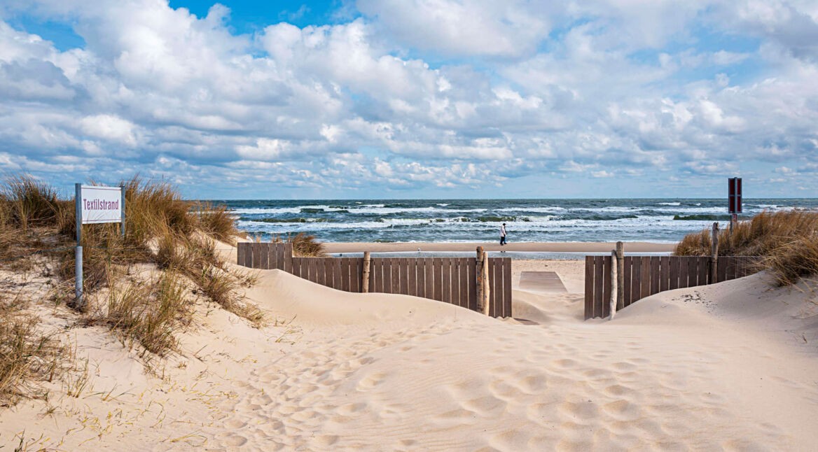 Ein Strandzugang zwischen den Dünen, im Hintergrund das Meer mit Schaumkronen
