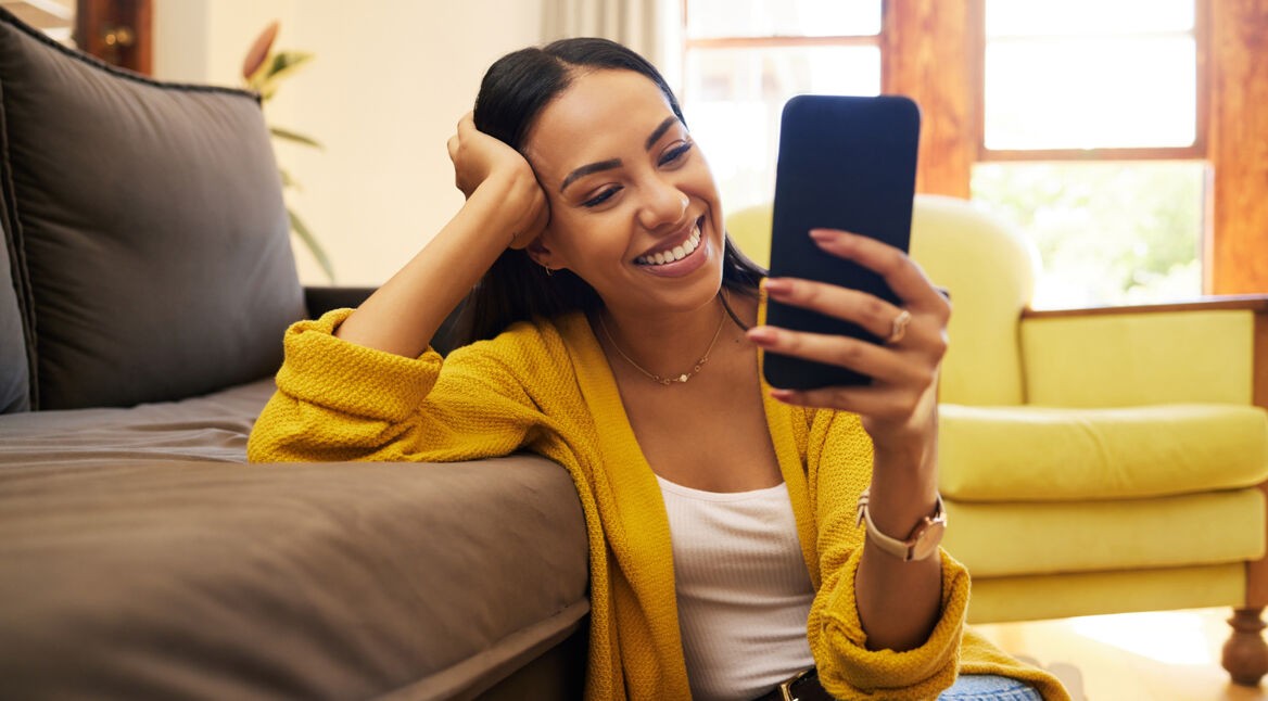Auf dem Bild zur Butterflies App sitzt eine Frau auf dem Boden vor einem Sofa und schaut lächelnd auf das Smartphone in ihrer Hand.