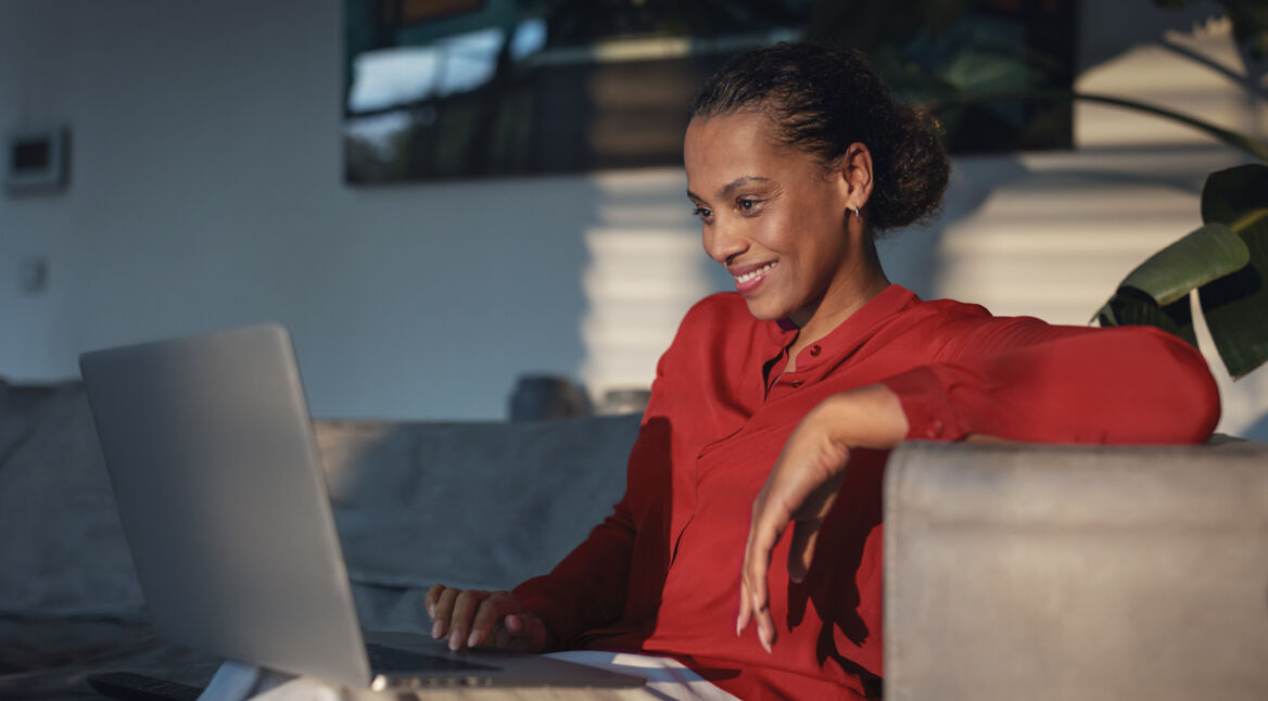 Auf dem Bild im Artikel zu Vodafone connect Festnetztest 2024 siehst Du eine Frau, die lächelnd in ihrem Wohnzimmer an ihrem Laptop sitzt. Sie trägt ein rotes Oberteil.